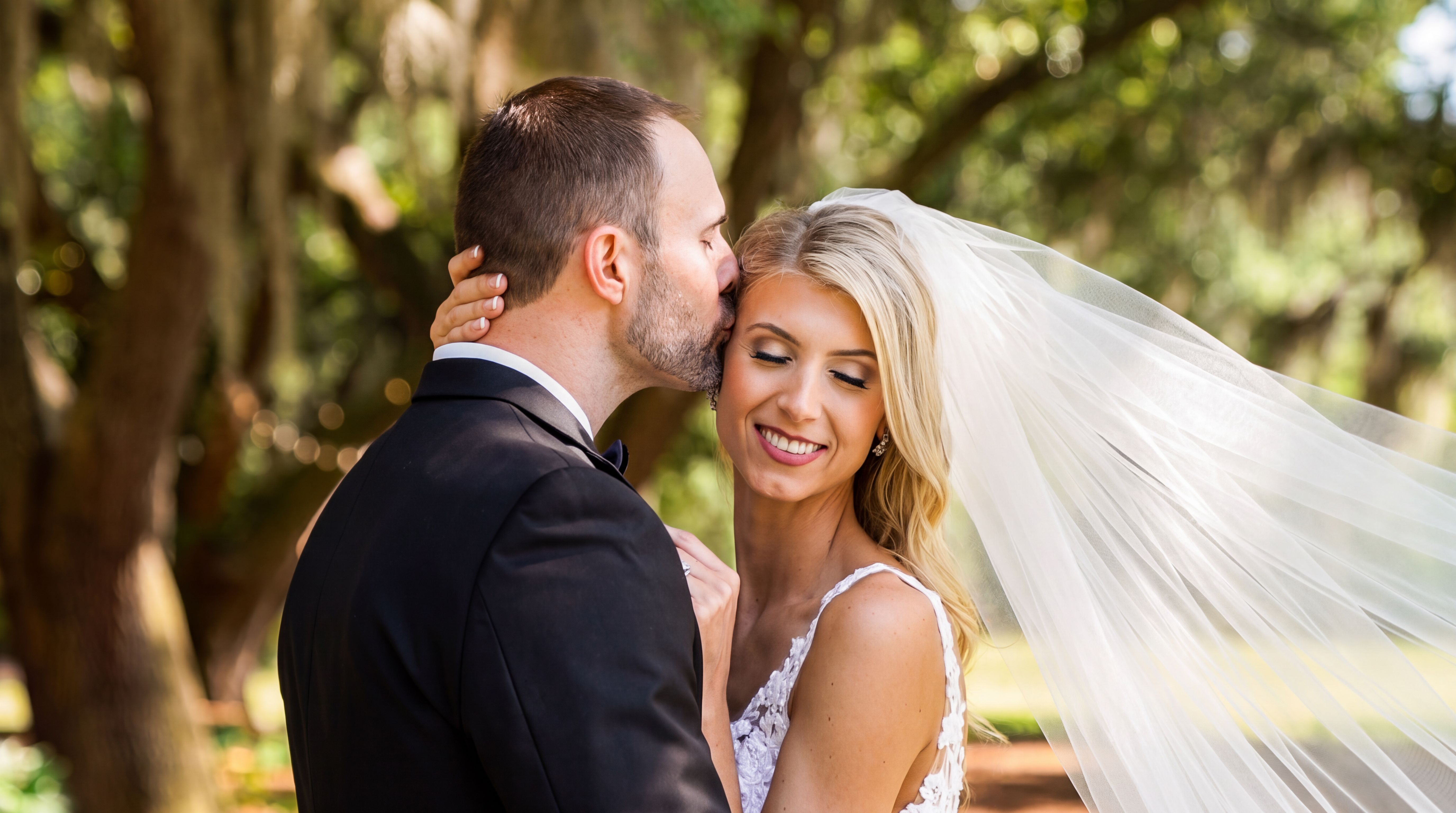 Groom kissing his bride beside her flowing veil during an outdoor wedding portrait