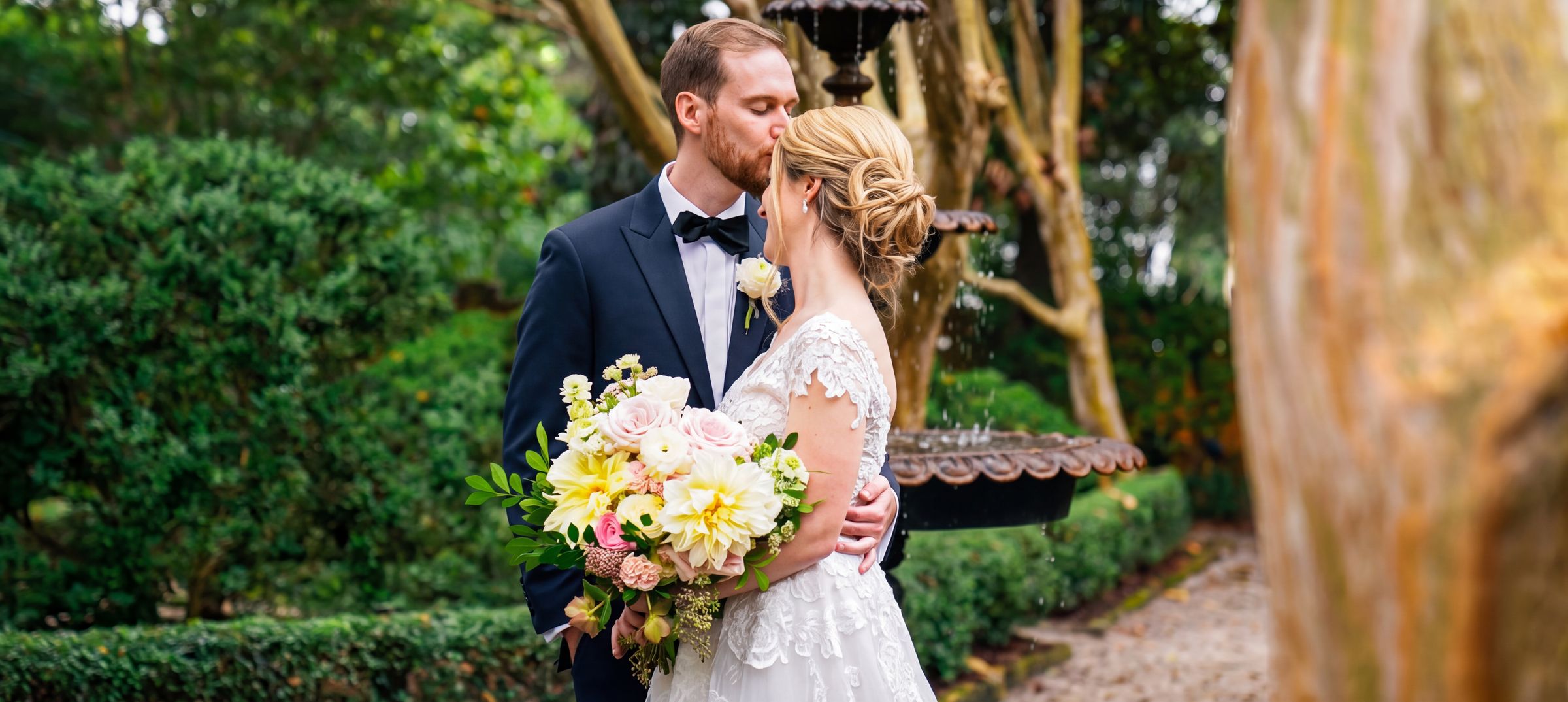Groom kissing his bride beside a garden fountain, holding a yellow bouquet