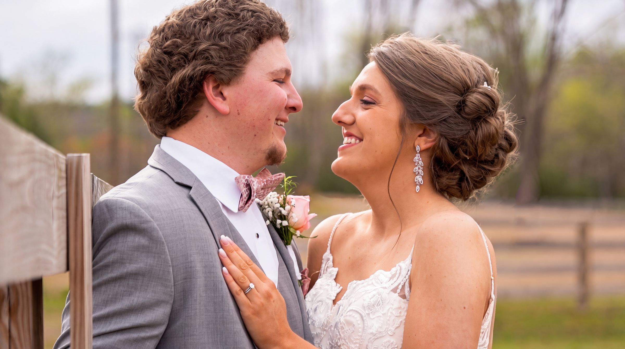 Groom in a gray suit and bow tie laughing with his bride beside a wooden fence
